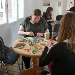 A group of women are sat around tables creating small pieces of art from textiles and found objects