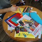 A man is sat at a table creating a zine from coloured paper, newspapers and pens